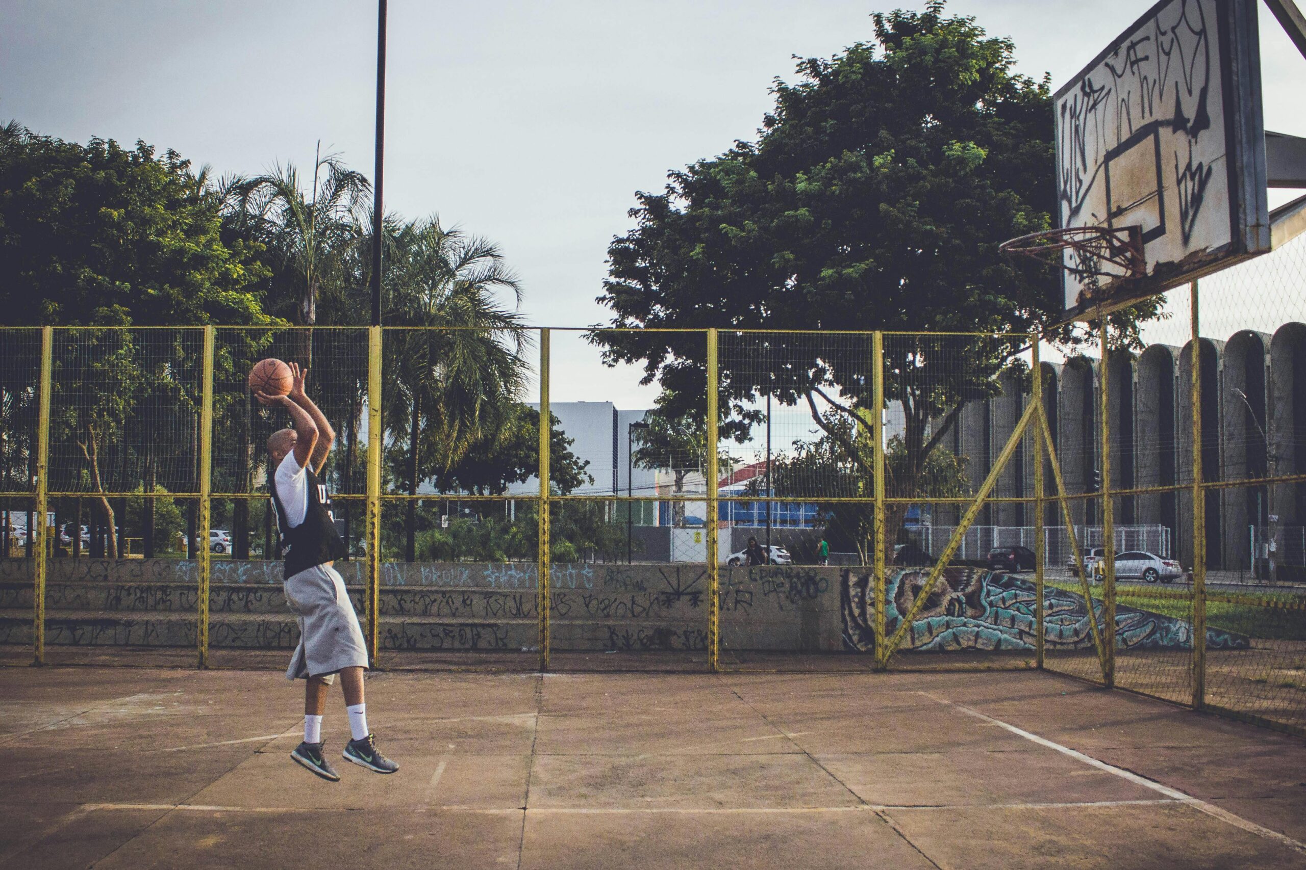 A man in athletic wear leaps for a basketball shot on an outdoor urban court.